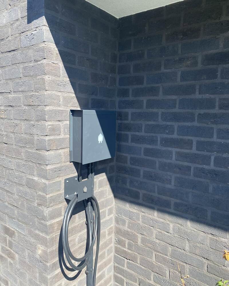 Borne de recharge murale gris anthracite, câble noir enroulé sur son support, contre un mur en briques.