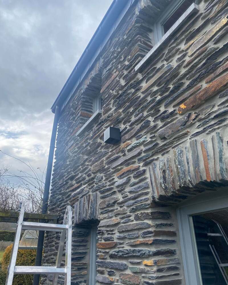 Façade en pierre d’une maison ardennaise avec une applique extérieure noire ; une échelle contre le mur.