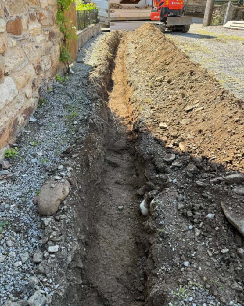 Tranchée creusée le long d’une maison en pierre pour la prise de terre ; une mini-pelle au fond de la cour.