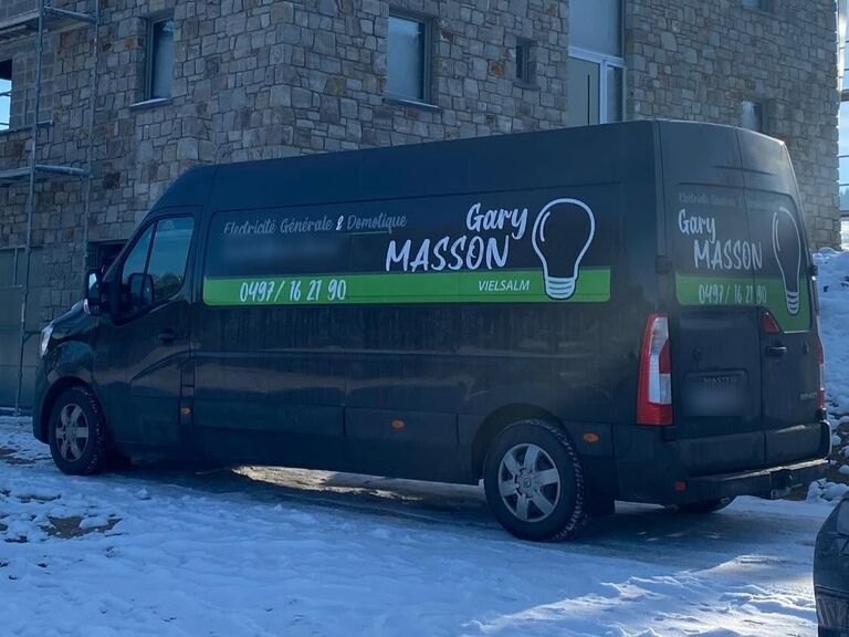 Camionnette noire lettrée « Gary Masson », ampoule et bandeau vert « Vielsalm », garée dans la neige devant une maison en pierre.