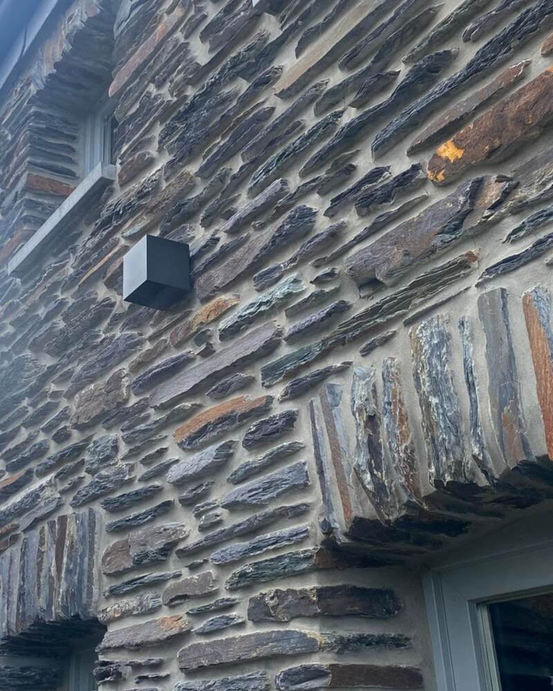 Façade en pierre d’une maison ardennaise vue de près : applique extérieure noire fixée au mur, près du linteau d’une fenêtre.