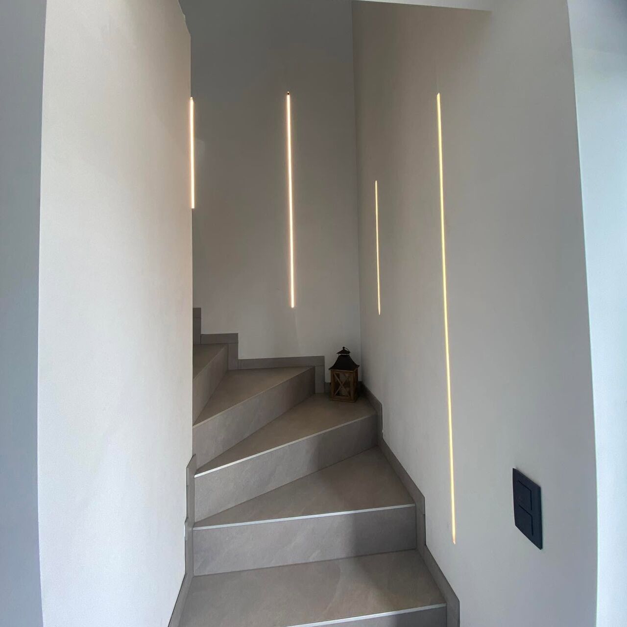 Cage d’escalier aux murs blancs éclairée par de fines lignes LED verticales encastrées ; interrupteur noir au mur.